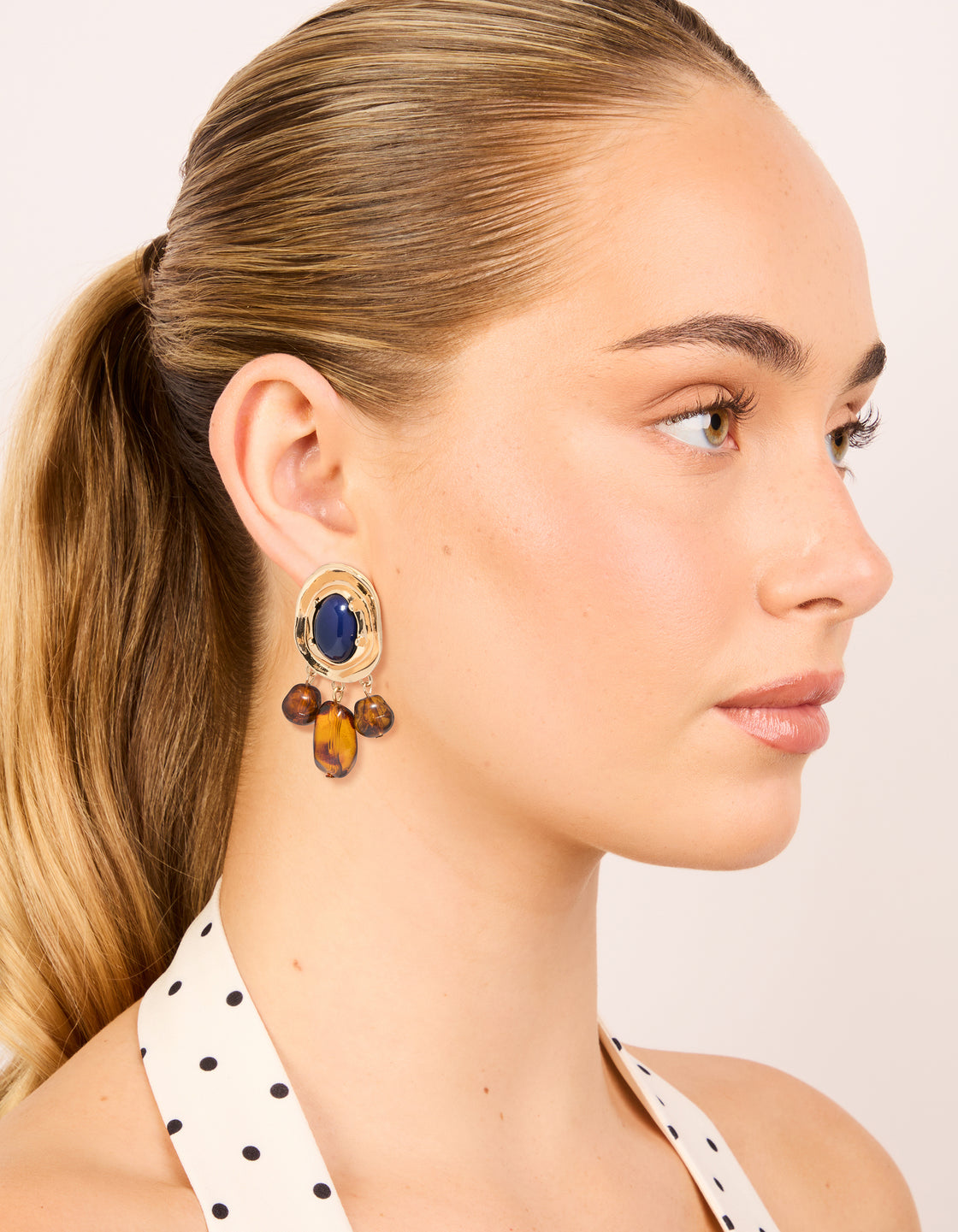 Displaying statement charm and beaded medallion drop earrings in gold with midnight blue acrylic and saddle brown accents on model view