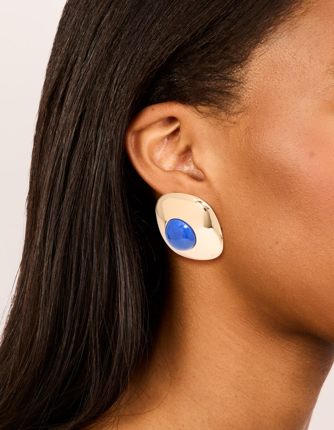 Showcasing statement stud earrings in gold with dark blue acrylic medallion in an on ear view
