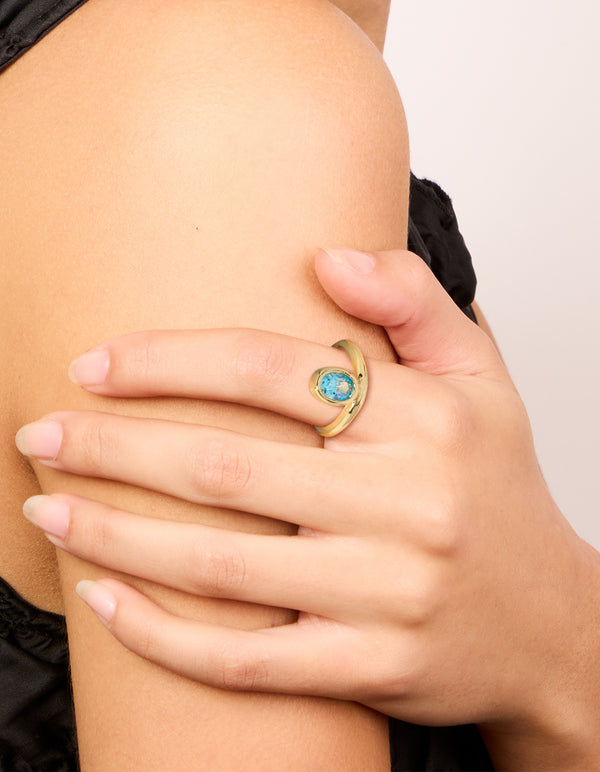 Featuring a gold plated stainless steel statement ring with oval cut blue cubic zirconia crystal in aqua on finger view